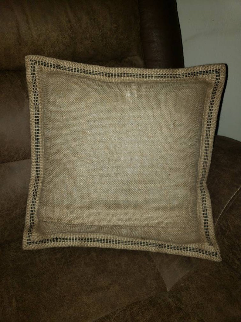 May include: A square burlap pillow with a black trim. The pillow is sitting on a brown leather couch.