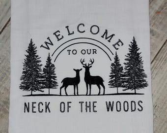 May include: White kitchen towel with black embroidery that reads "Welcome to our Neck of the Woods". The design features two deer and pine trees.