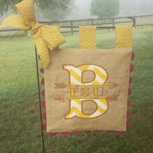 May include: A burlap garden flag with a large yellow and white chevron letter "B" with the text "FSU" in yellow on a red arrow. The flag has a scalloped edge and is attached to a black metal stake.