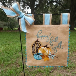 May include: A burlap garden flag with a light blue and tan bow. The flag features embroidered pumpkins and the words "happy fall y'all" in teal. The flag is trimmed with teal pom-poms.