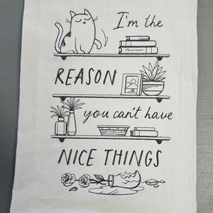 May include: Cream-coloured kitchen towel with a black sketched design. The design features a cat on a shelf, books, plants, and the text "I'm the REASON you can't have NICE THINGS."