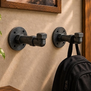 May include: Two wall-mounted hooks constructed from dark gray metal industrial piping. Each hook features a circular mounting flange with four screw holes, a short horizontal pipe segment, and an L-shaped end cap. A black backpack hangs from one hook.