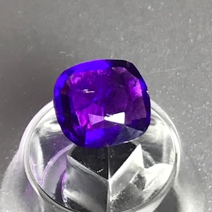 May include: A cushion-cut, deep purple gemstone with a faceted surface that reflects light. The stone is securely held in a clear, circular plastic display stand that features a central support structure to elevate the gem for viewing.