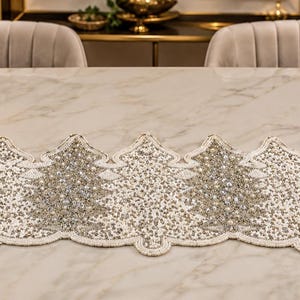 May include: A rectangular table runner featuring a repeating pattern of stylized Christmas trees. The runner is intricately decorated with silver and white beads and sequins, creating a shimmering texture. The edges follow the tree shapes.