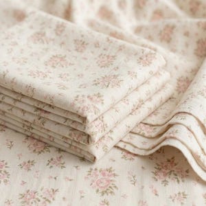 May include: A stack of folded fabric featuring a light beige background with a repeating pattern of small, soft pink floral clusters and delicate green leaves. The textile has a visible woven texture and is layered in multiple neat, horizontal folds.