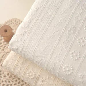 May include: Two folded pieces of white fabric featuring vertical stripes of intricate floral embroidery. The textile displays a repeating pattern of small blossoms and delicate lace-like detailing, creating a textured surface across the material.