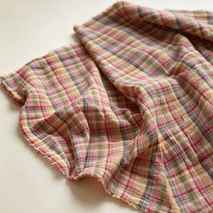 May include: A lightweight fabric featuring a multi-colored plaid pattern with intersecting lines in shades of red, yellow, green, and blue. The textile has a soft, crinkled texture and raw, frayed edges, showing a loose weave and semi-sheer quality.