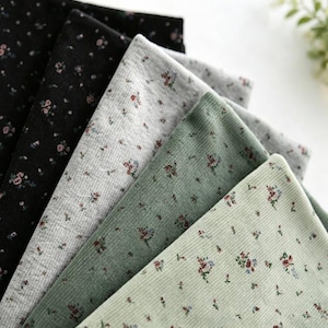 May include: A collection of folded ribbed fabric swatches featuring a small floral pattern. The colours include black, light grey, sage green, and pale mint green. Each piece of fabric displays a repeating print of tiny red and blue flowers.