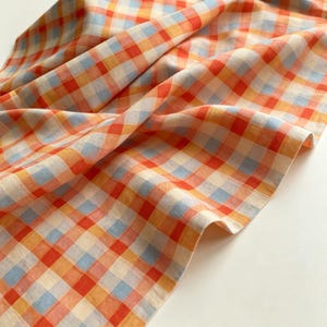 May include: A fabric textile featuring a repeating gingham check pattern in shades of bright orange, soft yellow, and light blue. The material is draped in soft folds, showcasing the consistent grid design across the woven surface of the cloth.