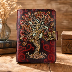 Puede incluir: Un diario de cuero rectangular con un acabado en rojo intenso que presenta un diseño de árbol en relieve con hojas doradas y naranjas. La cubierta está asegurada por un mecanismo de cierre de latón en el lado derecho, con intrincados patrones en espiral.