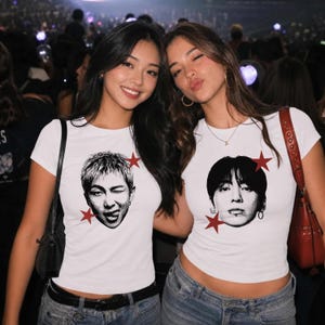 Op de afbeelding: Twee witte T-shirts met korte mouwen en een ronde hals, voorzien van zwart-wit halftoon portretgrafieken. Elk shirt is versierd met twee rode sterpictogrammen bij het portret. De shirts worden gedragen met een blauwe spijkerbroek.