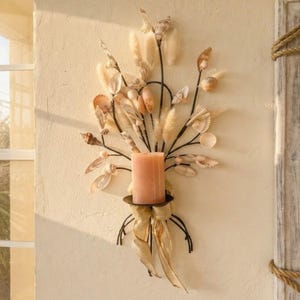 May include: A wall-mounted black metal candle holder featuring a cylindrical peach-colored pillar candle. The arrangement is decorated with various light-colored seashells and fluffy dried grasses, finished with a gold-toned fabric ribbon.
