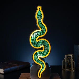 Op de afbeelding: Een verticale slangvormige lamp met een felgroen getextureerd lichaam en een oplichtende gele omlijning. De verlichte slang is gemonteerd op een donkere metalen voet, die op een rechthoekig houten platform staat voor een opvallend effect.