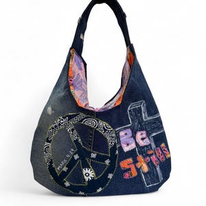 Peut inclure: Un sac hobo en jean bleu foncé orné d'un grand signe de paix appliqué avec le texte Mark 4:39 et d'une croix avec le texte Be Still. Le sac possède une bandoulière unique et une doublure en tissu à motifs colorés visible à l'ouverture.