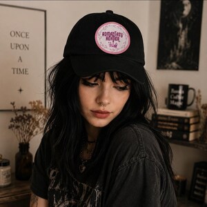 May include: A black baseball cap featuring a circular patch on the front. The patch has a light pink border with a white center, displaying the text ROMANTASY READER in a dark pink font, accompanied by a small pink crown graphic at the bottom.