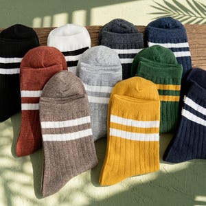 May include: A collection of ribbed crew socks in various colours including black, white, rust, grey, brown, mustard yellow, dark green, and navy blue. Each pair features two horizontal white or yellow stripes near the top cuff of the sock.