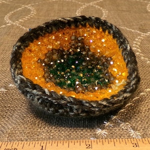May include: A small, round, crocheted bowl with a dark grey rim, a bright yellow middle section, and a dark green center filled with small, reflective beads. A wooden ruler placed in front shows the bowl is approximately 3.5 inches wide.