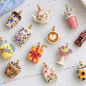 Könnte beinhalten: Eine Sammlung von zwölf Miniatur-Keramikanhängern mit verschiedenen Motiven, darunter eine Teekanne, eine Kaffeetasse mit Herz-Latte-Art, eine mit Loda beschriftete Sodaflasche, ein Geschenk, ein Blumen-Einmachglas, ein Teebeutel und Tassen.