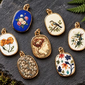 May include: Seven oval-shaped gold-rimmed pendants featuring diverse designs including a blue floral piece, a bird on a branch, orange marigolds, a portrait of a person, a hummingbird with white flowers, a paisley pattern, and a colorful floral motif.