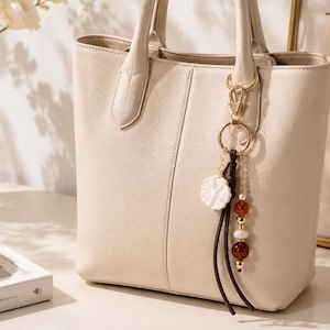 May include: A cream-colored leather tote bag with two top handles and a vertical seam down the center. A gold-toned metal keychain hangs from one handle, featuring a white flower charm, dark brown leather cords, and a strand of amber and pearl beads.