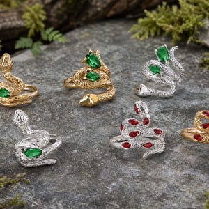 May include: A collection of six snake-shaped rings in gold and silver tones. Each ring features a textured, scaled metal body and is accented with pear-shaped, faceted gemstones in vibrant shades of emerald green or deep ruby red.