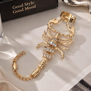 Könnte beinhalten: Ein goldfarbenes Handkettchen in Skorpionform mit klaren Strasssteinen, die entlang des Schwanzes und der Körpersegmente eingearbeitet sind. Das Schmuckstück besteht aus einem Ring oben und einem feinen Kettenarmband mit verstellbarem Verschluss.