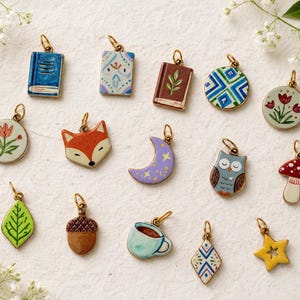 May include: A collection of fourteen small metal charms featuring various enamel designs including books, a fox, a crescent moon, an owl, a mushroom, a leaf, an acorn, a coffee cup, a star, and floral patterns, each with a gold-toned jump ring.
