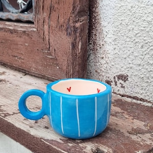 Könnte beinhalten: Eine kleine, runde, leuchtend blaue Keramiktasse mit vertikalen weißen Streifen auf der Außenseite. Das Innere der Tasse ist cremefarben und mit kleinen, roten, handgemalten Herzformen entlang des inneren Randes verziert.