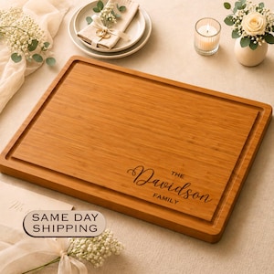 May include: A rectangular bamboo cutting board with a juice groove along the perimeter. The board features a laser-engraved inscription in the bottom right corner that reads "THE Davidson FAMILY" in a combination of script and serif fonts.