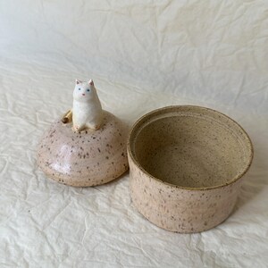 May include: A ceramic lidded jar with a speckled beige finish. The lid features a small, sculpted white cat figurine with blue eyes and a pink nose. The jar is cylindrical with a matching speckled texture, showing an open interior cavity.