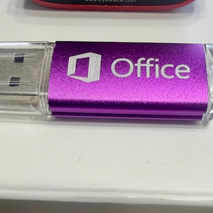 May include: A purple USB flash drive featuring a metallic finish and the white printed text Office with a corresponding logo. The device includes a clear plastic cap covering the metal connector and a small loop at the opposite end for attachment.