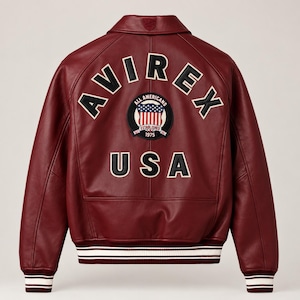 May include: A deep burgundy leather varsity jacket featuring a classic collar and ribbed cuffs with white stripes. The back displays the text AVIREX in large black letters, a circular All Americans 1975 patch with a flag motif, and USA below.