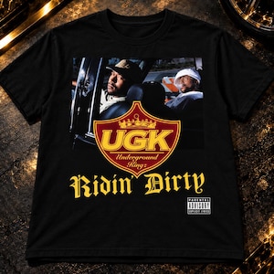 Puede incluir: Una camiseta negra de manga corta con un estampado gráfico de dos personas en un coche. Debajo de la imagen hay un escudo rojo y dorado con el texto UGK Underground Kingz, las palabras Ridin' Dirty en amarillo y una etiqueta de Parental Advisory.