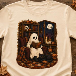 May include: A cream-colored t-shirt featuring a graphic of a ghost sitting in a chair reading a book. The scene includes a bookshelf with a skull, a black cat, pumpkins, candles, and a window showing a full moon and a bat in the night sky.