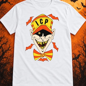 Puede incluir: Una camiseta blanca de manga corta con un gráfico de una cara de payaso que lleva una gorra amarilla y naranja con las letras ICP impresas en negro. El payaso tiene una amplia sonrisa con dientes amarillos, un parche negro y una pajarita.
