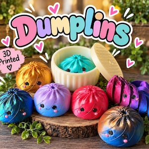 May include: A collection of 3D printed Dumplins figurines in various metallic and gradient colors including blue, gold, purple, red, and orange. Each round figurine features a small face with black eyes and pink cheeks. One blue Dumplin sits inside a small white container with a lid. The text Dumplins and 3D Printed are visible.