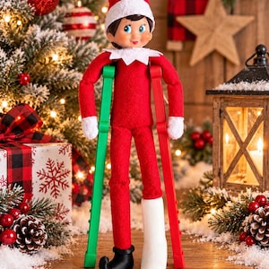 May include: A plush elf doll wearing a red suit with a white collar and hat, standing with one green crutch and one red crutch. The doll has a white cast on its left leg and a black shoe on its right foot, with a smiling face and blue eyes.