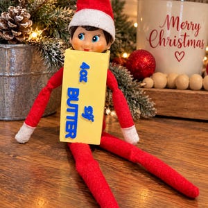 May include: A plush elf doll wearing a red suit and hat, holding a yellow rectangular block labeled 4oz BUTTER Elf in blue text. The doll has a smiling face with blue eyes and white cuffs on the sleeves. The block is positioned in front of the torso.