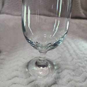 May include: A clear glass goblet with a smooth, rounded bowl, a short, sturdy stem, and a circular base. The glass is transparent and features a simple, functional design with a wide rim and a curved body that tapers down toward the stem.
