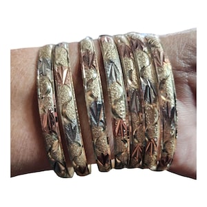 May include: A set of seven metallic bangles featuring a textured, sandblasted finish with alternating polished gold, silver, and rose gold tone accents. Each bangle displays a repeating etched pattern of curved lines and leaf-like shapes.
