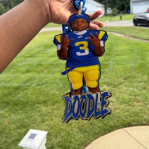 May include: A custom acrylic pendant featuring a photo of a football player wearing a blue jersey with the number 3 and yellow pants. The pendant includes the name DOODLE in blue and yellow lettering and is attached to a thick blue plastic chain.