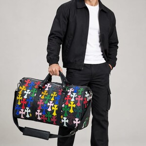 Puede incluir: Un bolso de viaje de cuero negro con un patrón repetitivo de parches en forma de cruz multicolor en rojo, amarillo, azul, verde y rosa. El bolso incluye dos asas superiores, una correa de hombro desmontable y herrajes de metal plateado.
