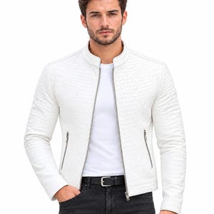 Puede incluir: Una chaqueta blanca con un patrón texturizado de relieve de cocodrilo, que presenta un cuello alzado, una cremallera frontal plateada de longitud completa y dos bolsillos laterales con cremallera. Se lleva sobre una camiseta blanca y vaqueros gris oscuro.