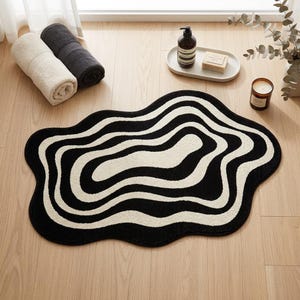 Peut inclure: Un tapis de bain de forme organique irrégulière présentant un motif contrasté de lignes ondulées et concentriques noires et crème. Le tapis possède une surface texturée douce et une silhouette fluide et arrondie rappelant des ondulations.