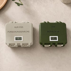 Könnte beinhalten: Zwei rechteckige Aufbewahrungsboxen mit Klappdeckeln und integrierten Digitalanzeigen. Die linke Box ist hellgrau mit dem Text "NUR FÜR FORSCHUNGSZWECKE", während die rechte Box olivgrün ist und den Aufdruck "TRT BIOHACKING" auf der Vorderseite trägt.
