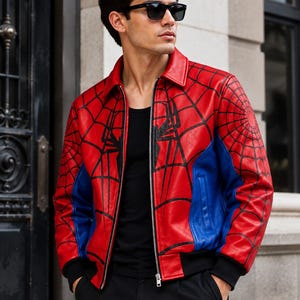 Op de afbeelding: Een rood en blauw leren jack met een zwart spinnenwebpatroon op de voorzijde en mouwen, een klassieke kraag, een zilveren ritssluiting en zwarte geribbelde boorden. Het jack wordt gedragen over een zwart hemd en een zwarte broek.