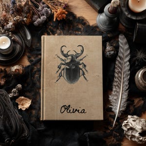May include: A tan hardcover book featuring a black ink illustration of a stag beetle centered on the front cover. Below the beetle, the name Olivia is printed in a black cursive script. The book has a smooth, matte-textured surface.