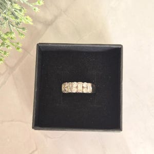 May include: A silver-toned ring featuring a row of alternating baguette and round-cut clear stones, centered within a square black velvet-lined jewelry box. The band has a metallic finish and the stones are set in a horizontal, linear arrangement.