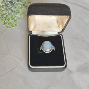 May include: A silver ring featuring an oval-shaped, milky white cabochon stone set in a serrated bezel. The ring is displayed inside a black velvet-lined jewellery box with a cream-coloured interior lid, presented in a professional manner.