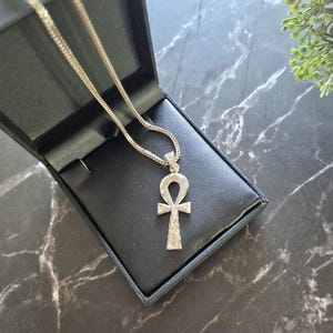 Op de afbeelding: Een zilverkleurige ankh-hanger ketting met een getextureerd, glinsterend oppervlak op het kruissymbool. De hanger hangt aan een bijpassende zilverkleurige ketting, gepresenteerd in een zwart juwelendoosje met een gladde, donkere binnenvoering.